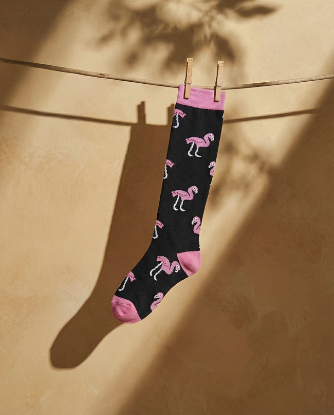 Black sock with pink flamingo pattern hanging on a clothesline against a beige wall.