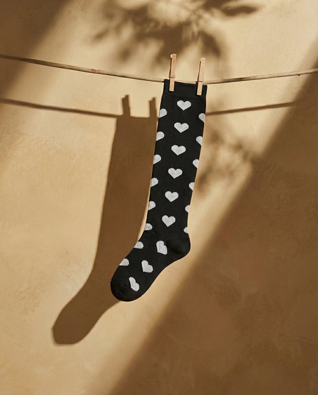 Black sock with white heart patterns hanging on a clothesline against a beige wall.