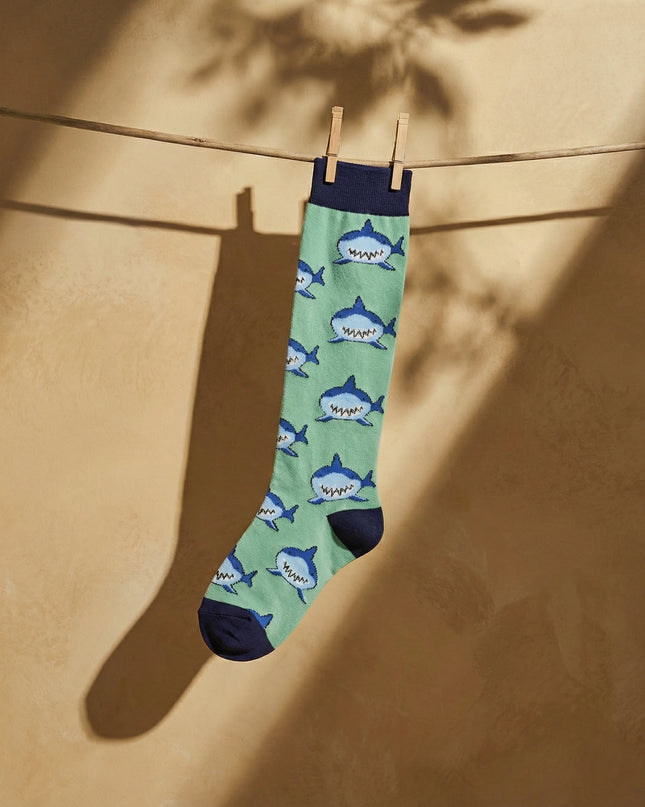 Green sock with shark pattern hanging on a clothesline against a beige wall.