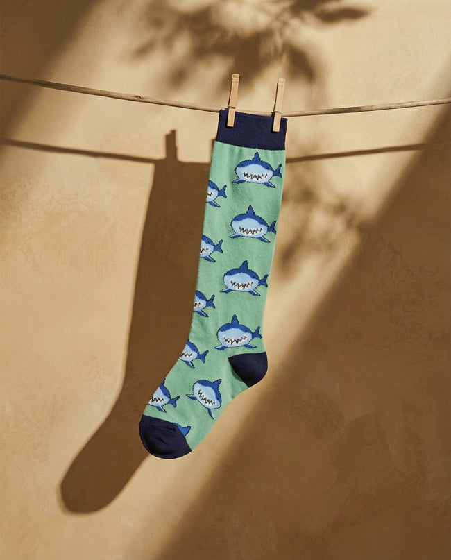 Green sock with shark pattern hanging on a clothesline against a beige wall.