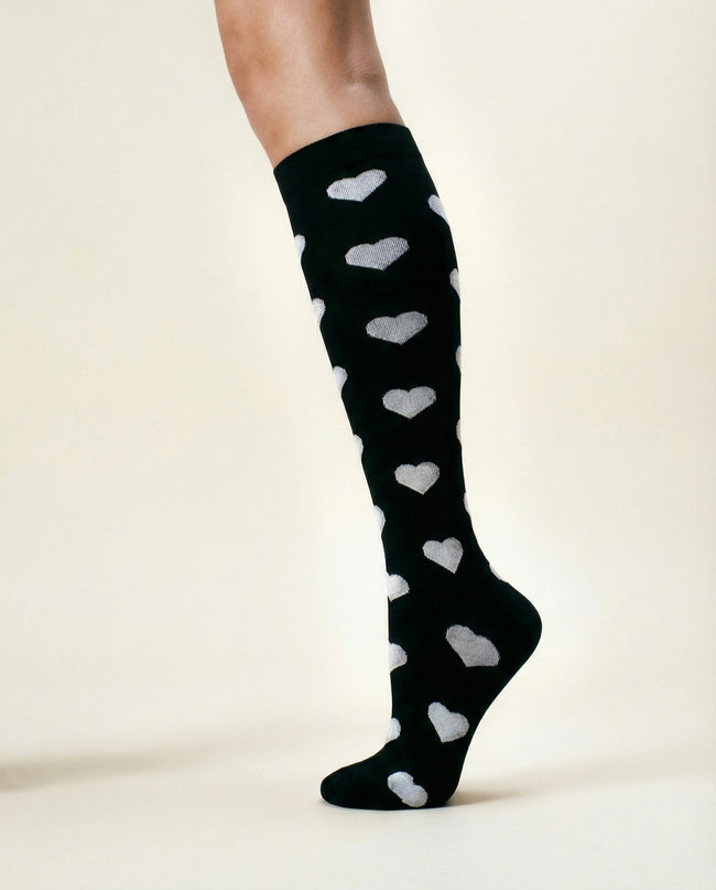 Black knee-high socks with white heart patterns worn by a person on a plain background