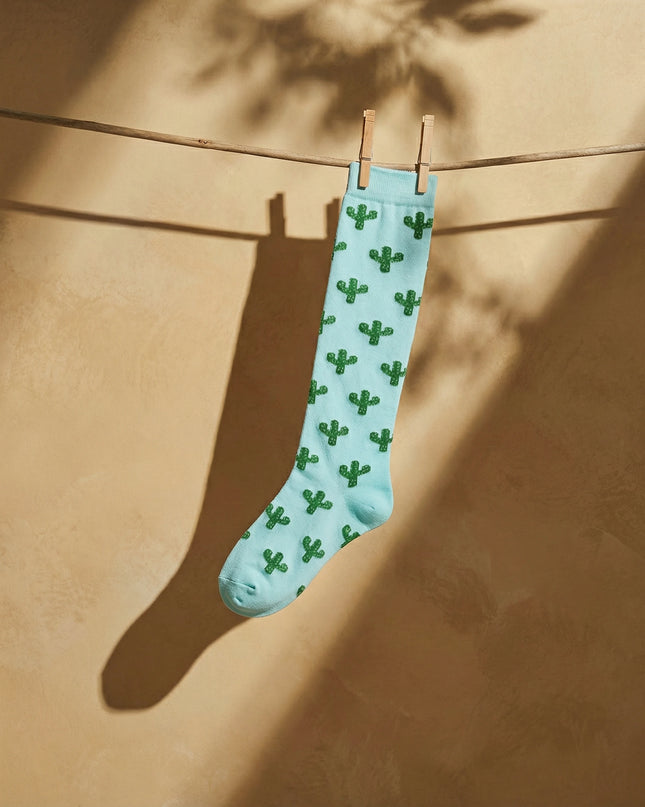 Light blue sock with green cactus pattern hanging on a clothesline against a beige wall.