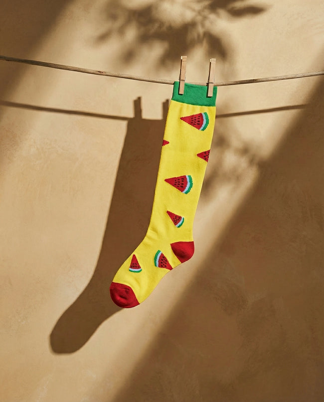 Yellow sock with watermelon pattern hanging on a clothesline against a beige wall.
