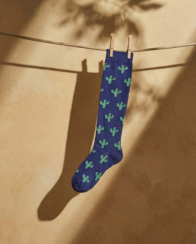 Blue sock with green cactus pattern hanging on a clothesline against a beige wall.