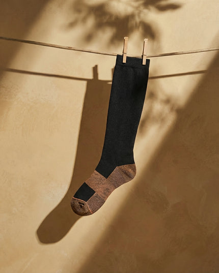 Black sock with brown sole hanging on a clothesline against a beige wall.