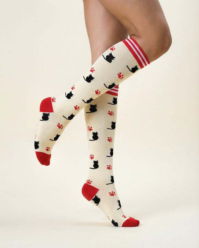 White knee-high socks with black cat and red paw print pattern worn by a person on a beige background