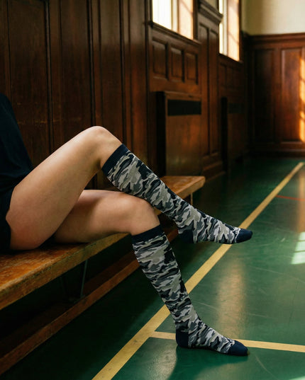 Person sitting on a wooden bench wearing camouflage-patterned knee-high socks in a sunlit hallway.