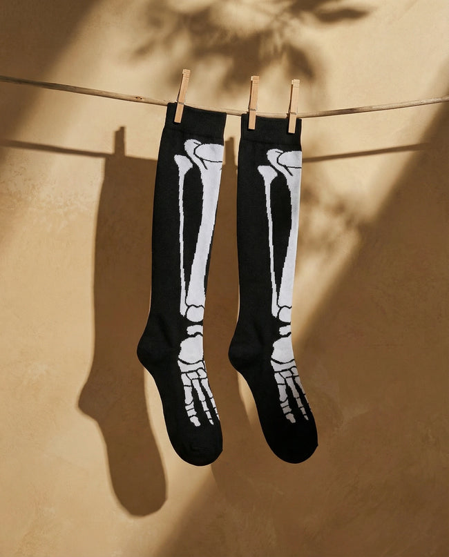 Black knee-high socks with white skeleton design hanging on a clothesline against a beige wall.