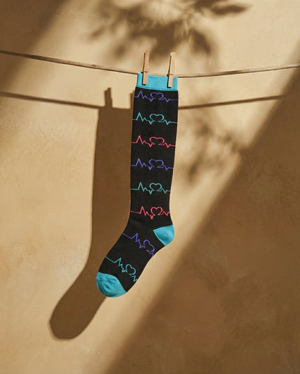 Black sock with colorful heart designs hanging on a clothesline against a beige wall.
